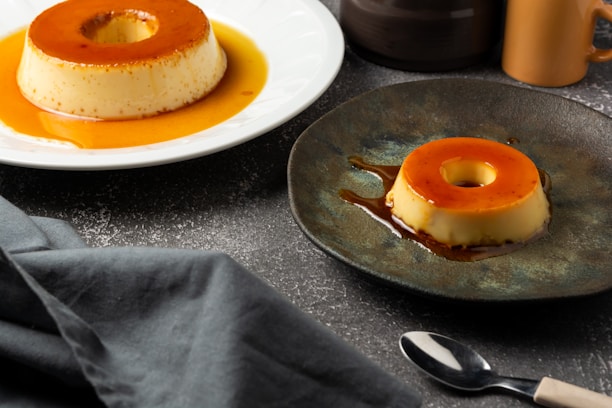 Two servings of caramel flan are presented, one on a white plate and the other on a dark, textured plate. The flans have a caramel glaze on top and are placed on a textured, dark-colored surface. Nearby are a black cloth, a small black spoon, and a ceramic cup.