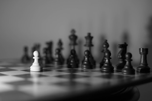 A black and white chessboard with a white pawn positioned prominently in the foreground, surrounded by various dark chess pieces in the background, giving a sense of focus and contrast.