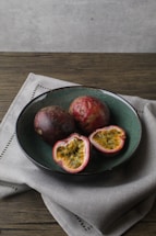 A teal-colored ceramic bowl contains three passion fruits, two whole and one sliced open to reveal the vibrant yellow pulp and black seeds. The bowl is placed on a neutral-toned fabric on a wooden surface with a soft, natural lighting backdrop.