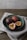 A teal-colored ceramic bowl contains three passion fruits, two whole and one sliced open to reveal the vibrant yellow pulp and black seeds. The bowl is placed on a neutral-toned fabric on a wooden surface with a soft, natural lighting backdrop.