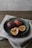 A teal-colored ceramic bowl contains three passion fruits, two whole and one sliced open to reveal the vibrant yellow pulp and black seeds. The bowl is placed on a neutral-toned fabric on a wooden surface with a soft, natural lighting backdrop.