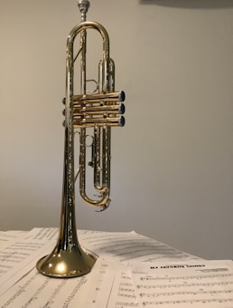A brass trumpet is standing upright on a pile of music sheets. The shiny surface of the trumpet reflects surrounding ambient light, and the music sheets are spread out beneath it, featuring various notes and a title that reads 'My Favorite Things'.