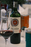A bottle of red wine labeled "Reserva Santa Ana" with a detailed circular design on the label. The bottle is accompanied by a wine glass partially filled with red wine. Other drink cans and bottles are visible in the background, placed on a white tablecloth. A plant with dark green leaves is slightly visible in the blurred background.