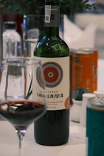 A bottle of red wine labeled "Reserva Santa Ana" with a detailed circular design on the label. The bottle is accompanied by a wine glass partially filled with red wine. Other drink cans and bottles are visible in the background, placed on a white tablecloth. A plant with dark green leaves is slightly visible in the blurred background.