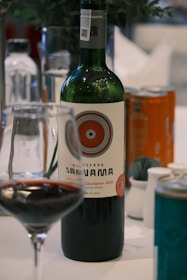 A bottle of red wine labeled "Reserva Santa Ana" with a detailed circular design on the label. The bottle is accompanied by a wine glass partially filled with red wine. Other drink cans and bottles are visible in the background, placed on a white tablecloth. A plant with dark green leaves is slightly visible in the blurred background.