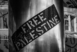 A monochrome image featuring a sticker with the words 'Free Palestine' placed on a metal surface. The background includes blurred architectural elements, possibly windows.