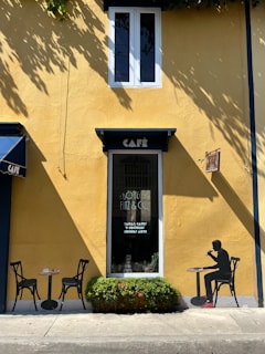 A small, charming café with a vivid yellow facade featuring a single window above and a door with a decorative sign. Two black chairs and a table are positioned outside next to some greenery. A silhouette of a person seated at a table on the wall adds an artistic touch.
