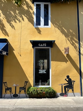 A small, charming café with a vivid yellow facade featuring a single window above and a door with a decorative sign. Two black chairs and a table are positioned outside next to some greenery. A silhouette of a person seated at a table on the wall adds an artistic touch.