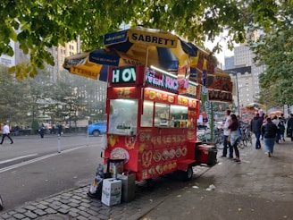 A vibrant street food cart is parked on a sidewalk under a canopy of green leaves. The cart is red with colorful signage displaying various food items and drinks. A couple of people are standing in front of the cart while others walk by on the street. Buildings and trees are visible in the background, creating an urban setting.