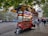 A vibrant street food cart is parked on a sidewalk under a canopy of green leaves. The cart is red with colorful signage displaying various food items and drinks. A couple of people are standing in front of the cart while others walk by on the street. Buildings and trees are visible in the background, creating an urban setting.