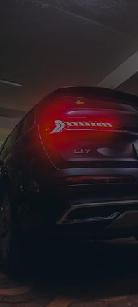 A dark, atmospheric image featuring the rear view of a car parked in an indoor space. The car's taillights are illuminated, casting a red glow. The model designation 'Q7' is visible on the vehicle.