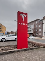 A large, prominent red sign with the Tesla logo and brand name is placed near a parking area. Surrounding the sign are parked cars, including a white Tesla Model S. The background features a street with buildings, including a brick building with multiple windows and a store with the Aldi logo. The sky appears overcast.