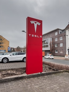 A large, prominent red sign with the Tesla logo and brand name is placed near a parking area. Surrounding the sign are parked cars, including a white Tesla Model S. The background features a street with buildings, including a brick building with multiple windows and a store with the Aldi logo. The sky appears overcast.