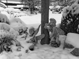 A winter scene featuring two fairy statues partially covered in snow. One fairy is sitting with wings and a playful expression, while the other is leaning against a tree, holding a leaf, with closed eyes. The ground and surrounding bushes are blanketed in a layer of fresh white snow, giving the area a peaceful and serene appearance. In the background, snow-covered plants and structures can be seen.