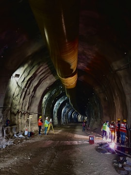 A tunnel under construction with workers present. The scene is dimly lit, revealing a long, cylindrical passageway with a large ventilation pipe running along the ceiling. Construction workers wearing helmets and safety vests are engaged in various activities. Some are inspecting the site with surveying equipment, while others appear to be managing or welding machinery.
