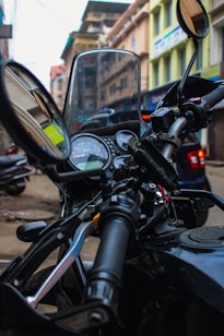 A close-up view of a motorcycle handlebar with visible control switches and mirrors, situated in an urban street setting. The background consists of blurred buildings and parked vehicles, indicating a city environment. The motorcycle dashboard is prominently shown with speedometer and other gauges.