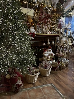 A festive room filled with Christmas decorations, including a brightly lit Christmas tree adorned with small white lights. Baskets filled with gold and red ornaments sit at the base, with Santa figurines placed nearby. The room is elaborately decorated with garlands, bells, and intricate holiday displays, creating a cozy and sumptuous atmosphere.