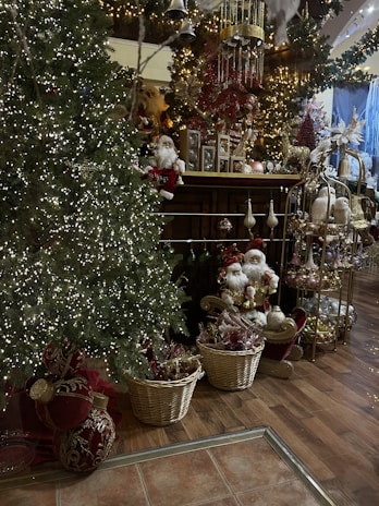 A festive room filled with Christmas decorations, including a brightly lit Christmas tree adorned with small white lights. Baskets filled with gold and red ornaments sit at the base, with Santa figurines placed nearby. The room is elaborately decorated with garlands, bells, and intricate holiday displays, creating a cozy and sumptuous atmosphere.
