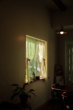A softly lit room showing a window with sheer curtains tied back, a small potted plant on the windowsill, and a chair partially visible near a table. The warm light creates a peaceful atmosphere. There is an overhead lamp hanging, and the corner of a painting or photograph resting against the wall.