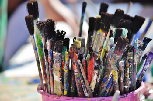 A collection of paintbrushes with varied colors and amounts of paint residue is placed in a pink container. The bristles are stained with multiple vibrant colors, suggesting the tools have been actively used in creative painting projects.