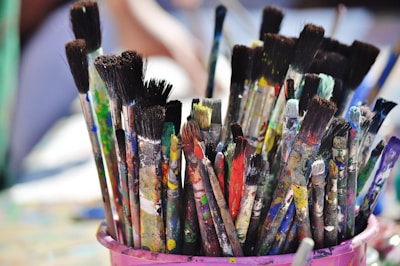 A collection of paintbrushes with varied colors and amounts of paint residue is placed in a pink container. The bristles are stained with multiple vibrant colors, suggesting the tools have been actively used in creative painting projects.