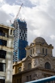 A construction site showing a skyscraper under development with scaffolding and a large crane on top. In the foreground, there is a historical stone building with ornate architecture and large windows, juxtaposed against the modern construction.