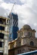 A construction site showing a skyscraper under development with scaffolding and a large crane on top. In the foreground, there is a historical stone building with ornate architecture and large windows, juxtaposed against the modern construction.