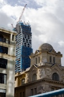 A construction site showing a skyscraper under development with scaffolding and a large crane on top. In the foreground, there is a historical stone building with ornate architecture and large windows, juxtaposed against the modern construction.
