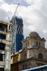 A construction site showing a skyscraper under development with scaffolding and a large crane on top. In the foreground, there is a historical stone building with ornate architecture and large windows, juxtaposed against the modern construction.