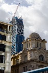A construction site showing a skyscraper under development with scaffolding and a large crane on top. In the foreground, there is a historical stone building with ornate architecture and large windows, juxtaposed against the modern construction.