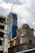 A construction site showing a skyscraper under development with scaffolding and a large crane on top. In the foreground, there is a historical stone building with ornate architecture and large windows, juxtaposed against the modern construction.