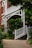 A charming exterior of a house featuring a red brick facade and white wooden railings. The entrance is adorned with lush green plants and trees, creating a welcoming and serene atmosphere. A curved pathway leads up to a set of steps that are lined with neatly trimmed bushes.