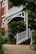 A charming exterior of a house featuring a red brick facade and white wooden railings. The entrance is adorned with lush green plants and trees, creating a welcoming and serene atmosphere. A curved pathway leads up to a set of steps that are lined with neatly trimmed bushes.