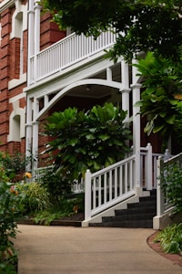 A charming exterior of a house featuring a red brick facade and white wooden railings. The entrance is adorned with lush green plants and trees, creating a welcoming and serene atmosphere. A curved pathway leads up to a set of steps that are lined with neatly trimmed bushes.
