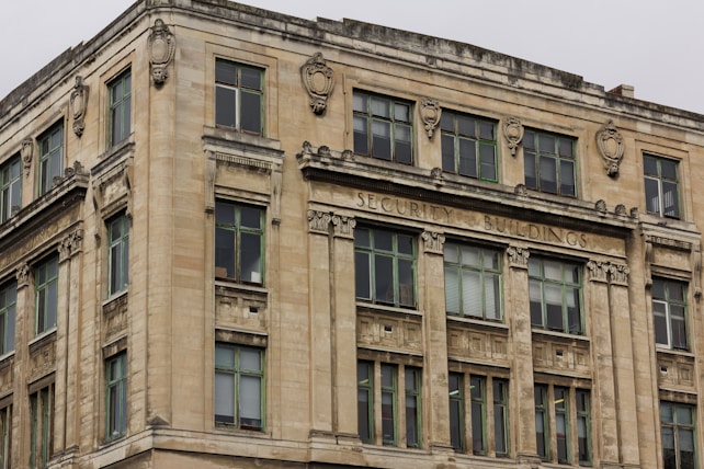 A large, historic building with a worn stone facade. The architecture features ornate embellishments around the windows and along the top section. The words 'Security Buildings' are prominently displayed across the top. The windows have dark frames and the overall color of the building is a weathered beige.