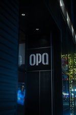 A dark, urban environment featuring a storefront with the sign 'OPA' illuminated against a dark wall. Yellow string lights are visible in the background, adding a festive element. Reflections of city lights in the glass create a vibrant, colorful atmosphere.