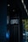 A dark, urban environment featuring a storefront with the sign 'OPA' illuminated against a dark wall. Yellow string lights are visible in the background, adding a festive element. Reflections of city lights in the glass create a vibrant, colorful atmosphere.