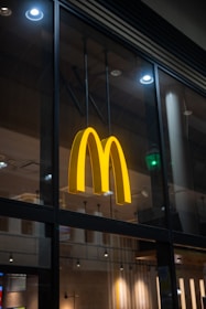 A large, illuminated yellow 'M' logo is mounted on the glass exterior of a building, possibly indicating a fast-food restaurant. The logo is prominent against a dark interior backdrop, with ceiling lights visible through the glass.