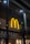 A large, illuminated yellow 'M' logo is mounted on the glass exterior of a building, possibly indicating a fast-food restaurant. The logo is prominent against a dark interior backdrop, with ceiling lights visible through the glass.