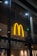 A large, illuminated yellow 'M' logo is mounted on the glass exterior of a building, possibly indicating a fast-food restaurant. The logo is prominent against a dark interior backdrop, with ceiling lights visible through the glass.
