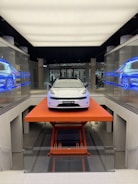 A sleek, modern white car is positioned on an orange lift platform in a futuristic, well-lit showroom. The showroom features glass walls with digital displays showing the car from different angles. The environment is clean and high-tech, enhancing the vehicle's sophisticated design.