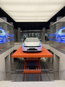 A sleek, modern white car is positioned on an orange lift platform in a futuristic, well-lit showroom. The showroom features glass walls with digital displays showing the car from different angles. The environment is clean and high-tech, enhancing the vehicle's sophisticated design.