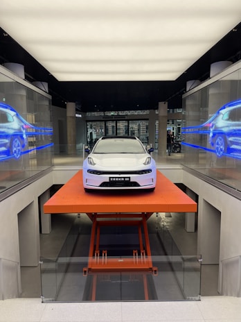 A sleek, modern white car is positioned on an orange lift platform in a futuristic, well-lit showroom. The showroom features glass walls with digital displays showing the car from different angles. The environment is clean and high-tech, enhancing the vehicle's sophisticated design.