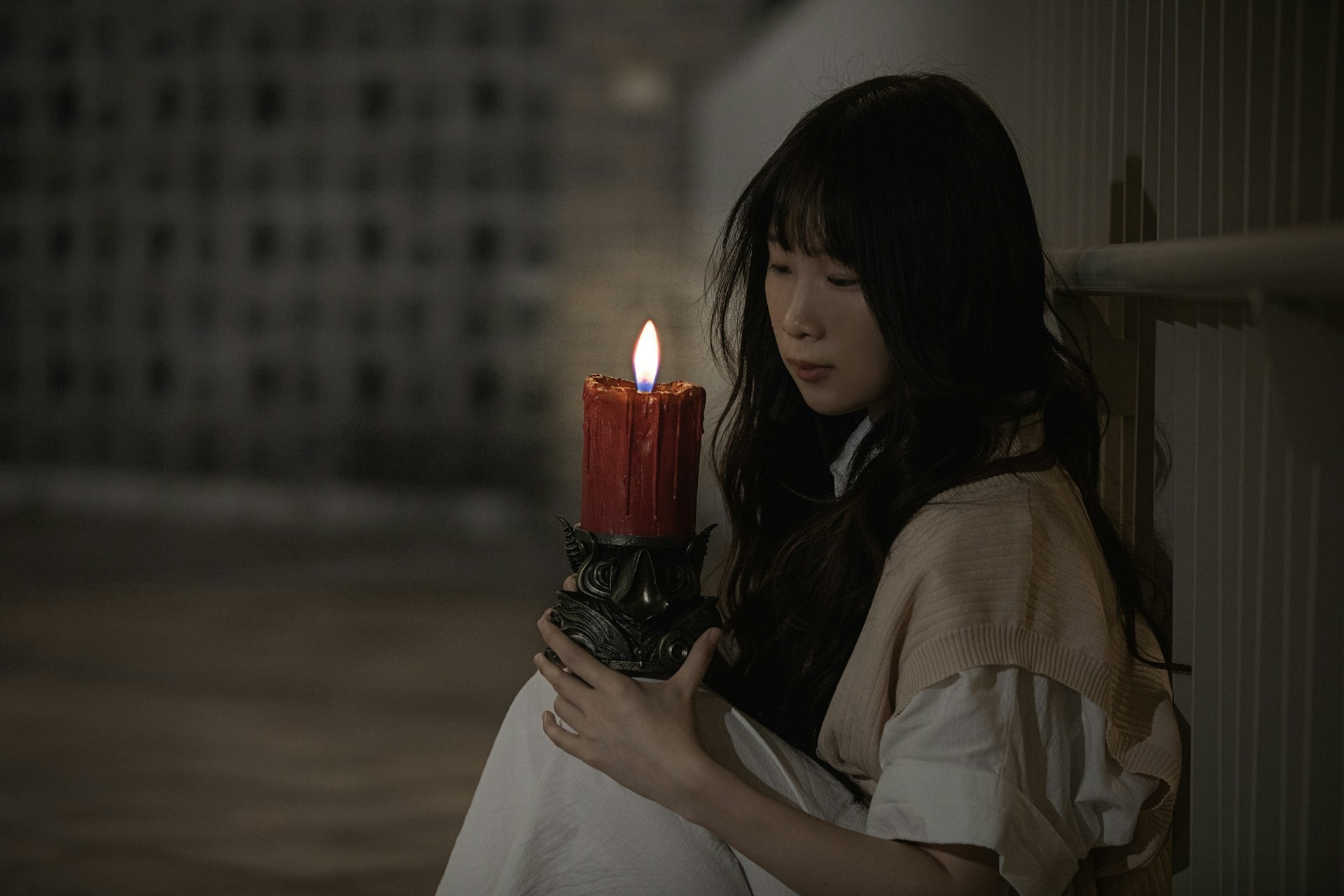 A young woman with long dark hair is sitting on the floor, holding a large, intricately designed candle with a reddish-orange flame. The surroundings are dimly lit, with a textured wall background and a railing on one side. The scene appears to convey a sense of solitude or contemplation.
