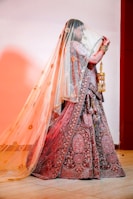 A person dressed in traditional, ornate clothing appears to be in a celebratory setting. The attire includes a richly embroidered skirt and a matching blouse with intricate patterns, complemented by detailed jewelry. A sheer veil with golden decorations is draped over the person, adding an elegant and festive touch. The person is holding golden accessories in their hand, which adds to the ornate appearance.