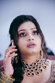 A woman with elaborate makeup and accessories holds a phone to her ear. She wears a heavy necklace adorned with red and gold accents and a matching intricate outfit. Henna designs decorate her hand.