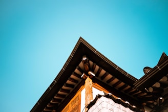 A traditional structure with a sharply angled roof against a clear blue sky. The roof features wooden beams and terracotta tiles, typical of East Asian architecture.