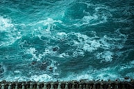 Turbulent ocean waves crash against a concrete breakwater, creating patterns of white foam on the deep teal water. A series of interlocking tetrapods line the shore, providing a stark contrast to the fluid movement of the sea.