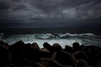 Dark clouds loom over a turbulent sea, with large waves crashing against a series of concrete tetrapods on the shore.
