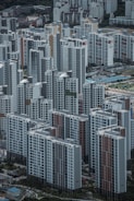 A densely packed urban landscape with numerous high-rise residential buildings. The structures are predominantly rectangular, featuring a mix of gray, white, and subtle earth tones. The perspective is from above, showcasing the organized grid layout typical of a cityscape. Parks and small roads can be seen between the buildings.